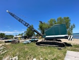 A large, green industrial crane positioned on a grassy area near a body of water. Several trees are in the background along with people standing near some parked vehicles and tents. The sky is clear and blue, indicating a sunny day.