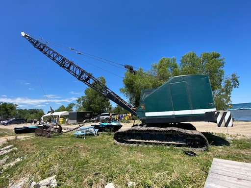 A large, green industrial crane positioned on a grassy area near a body of water. Several trees are in the background along with people standing near some parked vehicles and tents. The sky is clear and blue, indicating a sunny day.