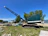 A large, green industrial crane positioned on a grassy area near a body of water. Several trees are in the background along with people standing near some parked vehicles and tents. The sky is clear and blue, indicating a sunny day.