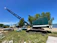 A large, green industrial crane positioned on a grassy area near a body of water. Several trees are in the background along with people standing near some parked vehicles and tents. The sky is clear and blue, indicating a sunny day.