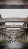 A view under a series of concrete overpasses, showing a road with vehicles including a van and motorcycles. The road is wet, suggesting recent rain. Signs on the left indicate directions to Surabaya and Pondok Taman, amidst greenery lining the road.