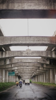 A view under a series of concrete overpasses, showing a road with vehicles including a van and motorcycles. The road is wet, suggesting recent rain. Signs on the left indicate directions to Surabaya and Pondok Taman, amidst greenery lining the road.