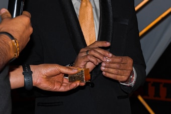 A person wearing a black suit and an orange tie is interacting with another individual holding a small golden box. The setting appears formal, and the focus is on the hands and the box. A microphone is also visible, suggesting a possible interview or presentation.