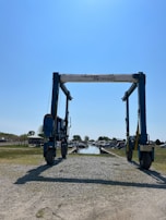 A large marine travel lift stands on a gravel path leading to a body of water. The machinery has a sturdy frame with wheels and is used for lifting boats. Surrounding the path are grassy areas and small buildings, with a clear blue sky overhead.