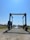 A large marine travel lift stands on a gravel path leading to a body of water. The machinery has a sturdy frame with wheels and is used for lifting boats. Surrounding the path are grassy areas and small buildings, with a clear blue sky overhead.