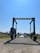 A large marine travel lift stands on a gravel path leading to a body of water. The machinery has a sturdy frame with wheels and is used for lifting boats. Surrounding the path are grassy areas and small buildings, with a clear blue sky overhead.