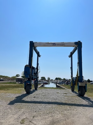 A large marine travel lift stands on a gravel path leading to a body of water. The machinery has a sturdy frame with wheels and is used for lifting boats. Surrounding the path are grassy areas and small buildings, with a clear blue sky overhead.