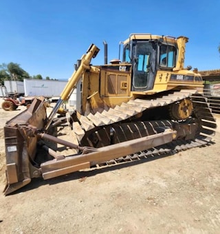 A large track-type bulldozer is parked on a dirt surface. It features a wide blade at the front used for pushing material and heavy tracks that allow it to move over rough terrain. The cab is positioned towards the back, with visible windows and control areas.