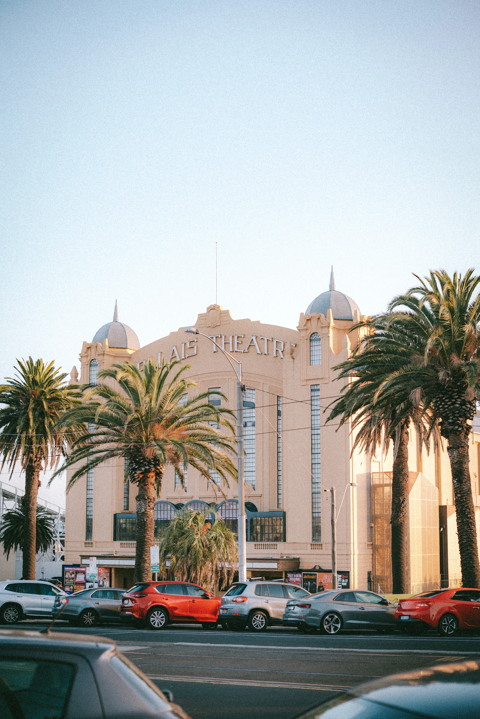 The historic Palais Theatre building with domed towers, palm trees, cars, and street traffic outside.