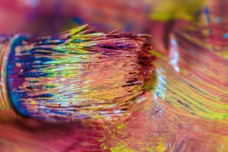 Close-up of a paintbrush with bristles coated in vibrant, multicolored paint resting on a surface covered in the same hues. The colors blend together, creating a dynamic spectrum with textures that suggest movement and creativity.