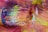 Close-up of a paintbrush with bristles coated in vibrant, multicolored paint resting on a surface covered in the same hues. The colors blend together, creating a dynamic spectrum with textures that suggest movement and creativity.