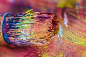 Close-up of a paintbrush with bristles coated in vibrant, multicolored paint resting on a surface covered in the same hues. The colors blend together, creating a dynamic spectrum with textures that suggest movement and creativity.