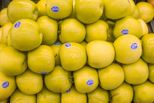 A large pile of yellow apples, each with a small blue sticker attached, is stacked together. The apples appear fresh and shiny, making them visually appealing.