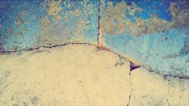 A cracked concrete surface with a textured, weathered appearance. The upper portion of the concrete is painted with a blend of blue and yellow, creating a somewhat rustic and aged look. The cracks run diagonally, with some faint discoloration around them.