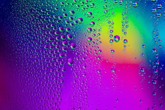 Droplets of water clinging to a surface, set against a vivid, multicolored background. The colors blend in gradients, with prominent hues of blue, purple, green, and pink. The water droplets are scattered across the surface, varying in size and giving a sense of texture.