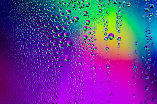 Droplets of water clinging to a surface, set against a vivid, multicolored background. The colors blend in gradients, with prominent hues of blue, purple, green, and pink. The water droplets are scattered across the surface, varying in size and giving a sense of texture.