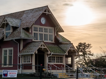 A two-story house with a steeply pitched roof and dormer windows. The facade is brown with white trim and features a clock above the upper windows. The roof is made of wooden shingles. Nearby, there's a construction sign, and the scene is illuminated by a bright sun in a partly cloudy sky. Trees and a fenced area are visible in the surroundings.