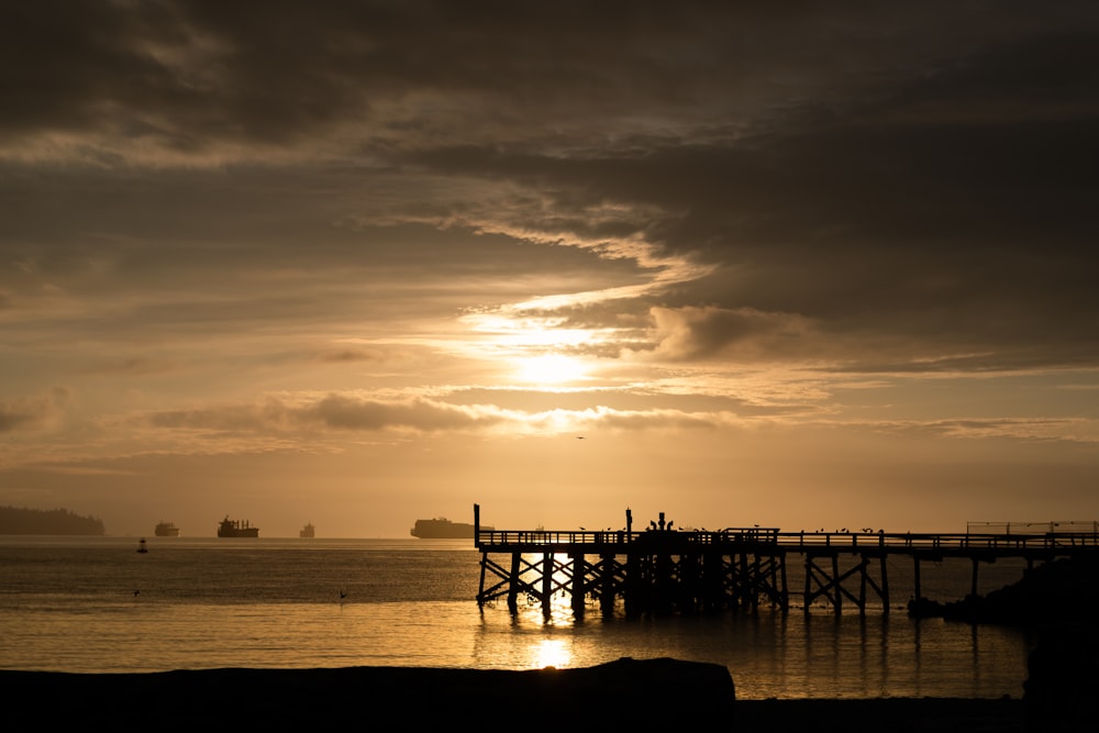the sun is setting over the ocean with a pier in the foreground