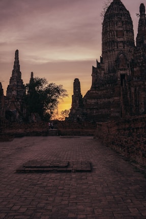Historic temple structures silhouetted against a dramatic sunset sky, with intricate architectural details visible on the ruins. The warm glow of the sunset illuminates the ancient bricks, creating a serene and nostalgic atmosphere.