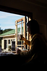 A serene morning scene with a woman journaling by a sunlit window, surrounded by soft greenery and a cup of tea.