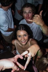 A group of young people enjoying themselves, with one person making a shaka hand gesture. The focus is on a woman smiling up at the camera, with her hand forming a heart shape with another hand. The setting appears to be a social gathering or party.