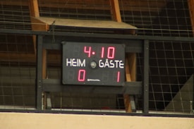 A digital scoreboard is mounted on a wall, displaying the time 4:10 and scores with the words 'HEIM' and 'GÄSTE', translating to 'Home' and 'Guests'. The home team has a score of 0, while the guest team leads with a score of 1.