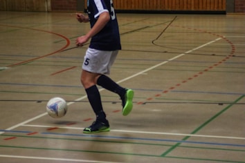 A person in a navy blue and white sports uniform is playing futsal in an indoor court. The individual is about to kick a colorful ball while wearing bright green and black shoes. The court is marked with various colored lines and has a wooden wall in the background.