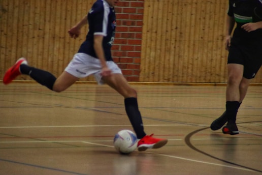 Two individuals are playing futsal in an indoor setting with a wooden and brick wall background. One player, wearing a dark jersey and bright red shoes, is about to kick a white soccer ball as the other player approaches.