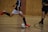 Two individuals are playing futsal in an indoor setting with a wooden and brick wall background. One player, wearing a dark jersey and bright red shoes, is about to kick a white soccer ball as the other player approaches.