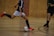 Two individuals are playing futsal in an indoor setting with a wooden and brick wall background. One player, wearing a dark jersey and bright red shoes, is about to kick a white soccer ball as the other player approaches.