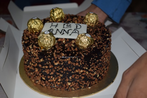A chocolate cake adorned with chocolate nut toppings and several gold-wrapped chocolates on top. A white chocolate plaque with 'HBD ANNA' written on it is centrally placed. The cake rests on a white surface, and a hand is partially visible along the edge, suggesting someone is handling the cake.