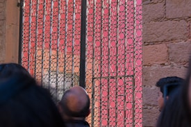 A decorative gate with intricate patterns made of black metal set against a pink background. The gate's design features a series of circular and abstract forms. Part of a stone wall is visible on the right side, and several people are partially seen in the foreground.