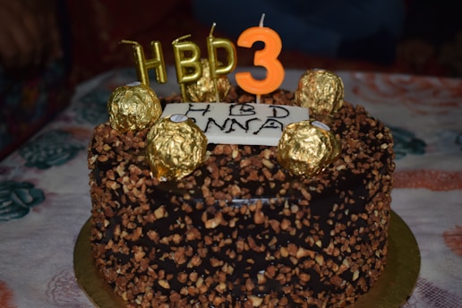 A chocolate cake decorated with nuts and gold-wrapped chocolates sits on a table. On top, candles spell out 'HBD' and the number '3'. A message is written on a white frosting plaque.