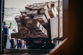 A truck is heavily loaded with numerous cardboard boxes, with some hanging precariously off the side. Several people are gathered around, one of whom is taking a photo with a phone. They appear to be in a warehouse or loading dock, under natural daylight.