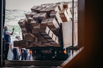 A truck is heavily loaded with numerous cardboard boxes, with some hanging precariously off the side. Several people are gathered around, one of whom is taking a photo with a phone. They appear to be in a warehouse or loading dock, under natural daylight.