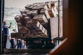 A truck is heavily loaded with numerous cardboard boxes, with some hanging precariously off the side. Several people are gathered around, one of whom is taking a photo with a phone. They appear to be in a warehouse or loading dock, under natural daylight.