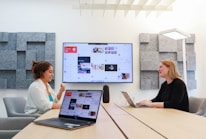 Two people are sitting across a conference table, each with a laptop open in front of them. A large screen displaying a digital interface with various content is mounted on the wall between them. The room has soundproofing panels on the walls and modern lighting.