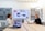 Two people are sitting across a conference table, each with a laptop open in front of them. A large screen displaying a digital interface with various content is mounted on the wall between them. The room has soundproofing panels on the walls and modern lighting.