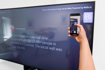 A person is holding a smartphone to take a picture of a large screen displaying a social media post. The content on the screen talks about increasing real-time engagement at a large event called GearFest. In the top corner, there's a QR code and a prompt to create a post.