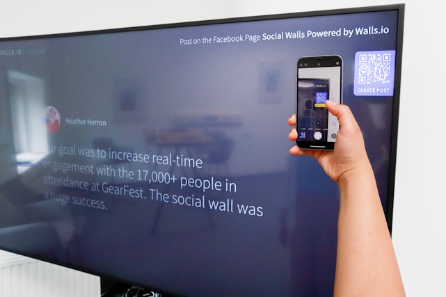 A person is holding a smartphone to take a picture of a large screen displaying a social media post. The content on the screen talks about increasing real-time engagement at a large event called GearFest. In the top corner, there's a QR code and a prompt to create a post.