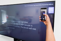 A person is holding a smartphone to take a picture of a large screen displaying a social media post. The content on the screen talks about increasing real-time engagement at a large event called GearFest. In the top corner, there
