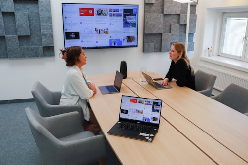Two people are sitting at a conference table in an office meeting room, each with a laptop in front of them. The room has a large screen mounted on the wall displaying a digital interface, and acoustic panels are visible on the walls. There is a window providing natural light, and the table is surrounded by gray chairs. A glass of water and a speaker are also present on the table.