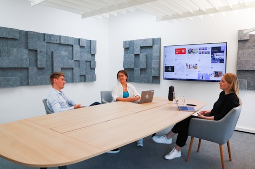 Three people are seated around a modern conference table in a well-lit meeting room. One person is using a laptop, another has a laptop closed in front of them, and the third is listening attentively. A large screen on the wall displays various media and web pages. The room has acoustic panels on the walls and a minimalist design.