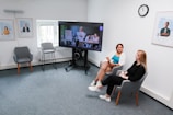 Two people sitting in a modern office space, engaged in conversation. A large monitor between them displays a video call featuring several participants. The room features a carpeted floor, a clock on the wall, and framed informational posters with a bright window.