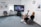 Two people sitting in a modern office space, engaged in conversation. A large monitor between them displays a video call featuring several participants. The room features a carpeted floor, a clock on the wall, and framed informational posters with a bright window.