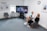 Two people sitting in a modern office space, engaged in conversation. A large monitor between them displays a video call featuring several participants. The room features a carpeted floor, a clock on the wall, and framed informational posters with a bright window.