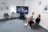 Two people sitting in a modern office space, engaged in conversation. A large monitor between them displays a video call featuring several participants. The room features a carpeted floor, a clock on the wall, and framed informational posters with a bright window.