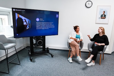 Two people are seated in comfortable chairs in an office setting, engaged in conversation. A large screen nearby displays information about a social media platform. There is a clock on the wall and a framed poster next to it. The room has modern decor with neutral tones.