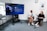 Two people are seated in comfortable chairs in an office setting, engaged in conversation. A large screen nearby displays information about a social media platform. There is a clock on the wall and a framed poster next to it. The room has modern decor with neutral tones.