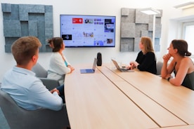 Four people are seated around a conference table in a modern office setting. A large screen on the wall displays various content, suggesting a presentation or discussion. The room is well-lit, with acoustic panels on the walls, and two laptops are open on the table.